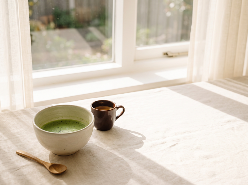 A bowl of bright green matcha beside a small cup of coffee on cream linen in morning window light