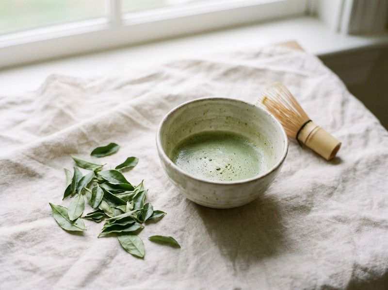 A bowl of whisked matcha with fresh tea leaves scattered beside it